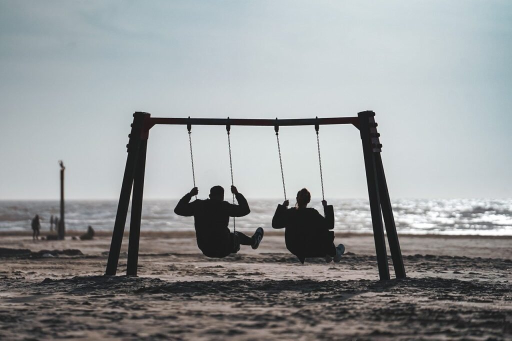 Beach Swing Silhouette: Two people sitting on a swing set by the beach, silhouetted against the soft, misty background of the ocean. The scene feels calm and serene, with a nostalgic, peaceful vibe.