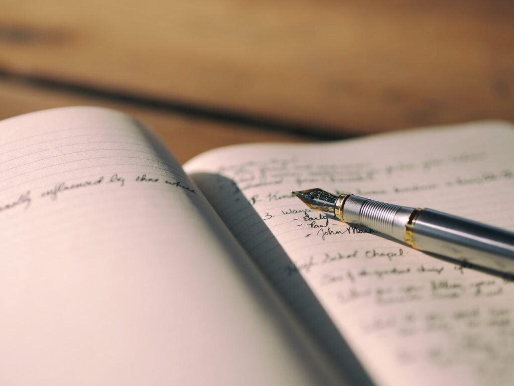 Open Notebook with Pen: An open notebook lying on a wooden surface with a black pen resting on it. The background is softly blurred, focusing on the writing and note-taking scene.