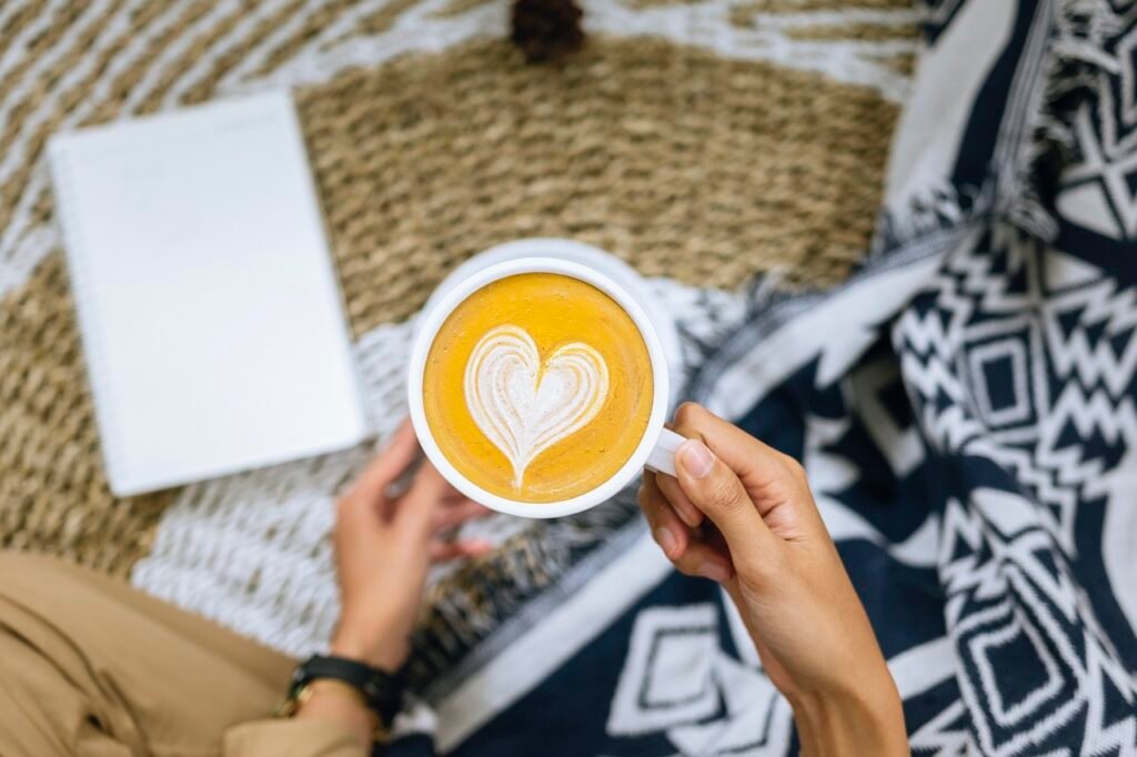 Self-Care Moment with Coffee: A person holding a cup of coffee with a heart-shaped foam design, sitting on a cozy blanket outdoors. There's a book and a tablet on the blanket, suggesting a relaxing self-care or me-time moment.