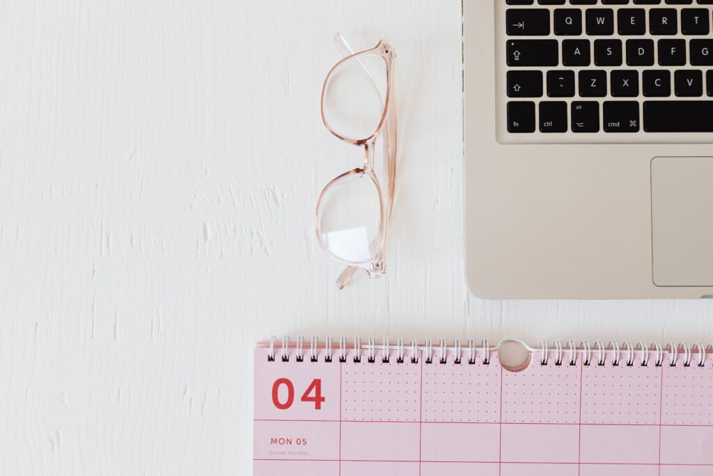 Top view of a pink calendar with a Monday date marked as April 5, glasses, and a laptop on a white desk