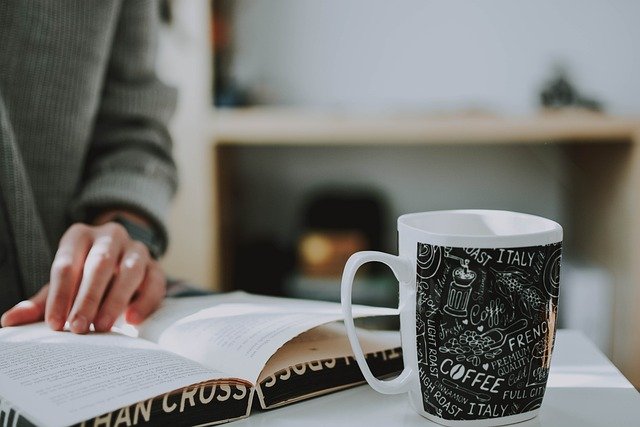 A cozy scene of a person reading a book with a black coffee mug decorated with text and illustrations, representing relaxation and a peaceful Sunday reset.